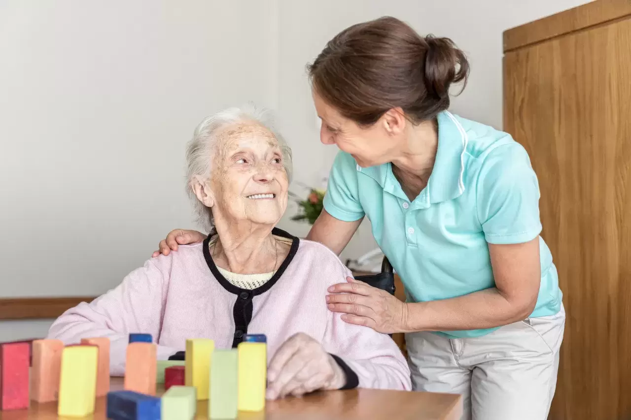 Pflegehelfende betreut eine ältere, lächelnde Dame, die sich mit Farbklötzen beschäftigt..