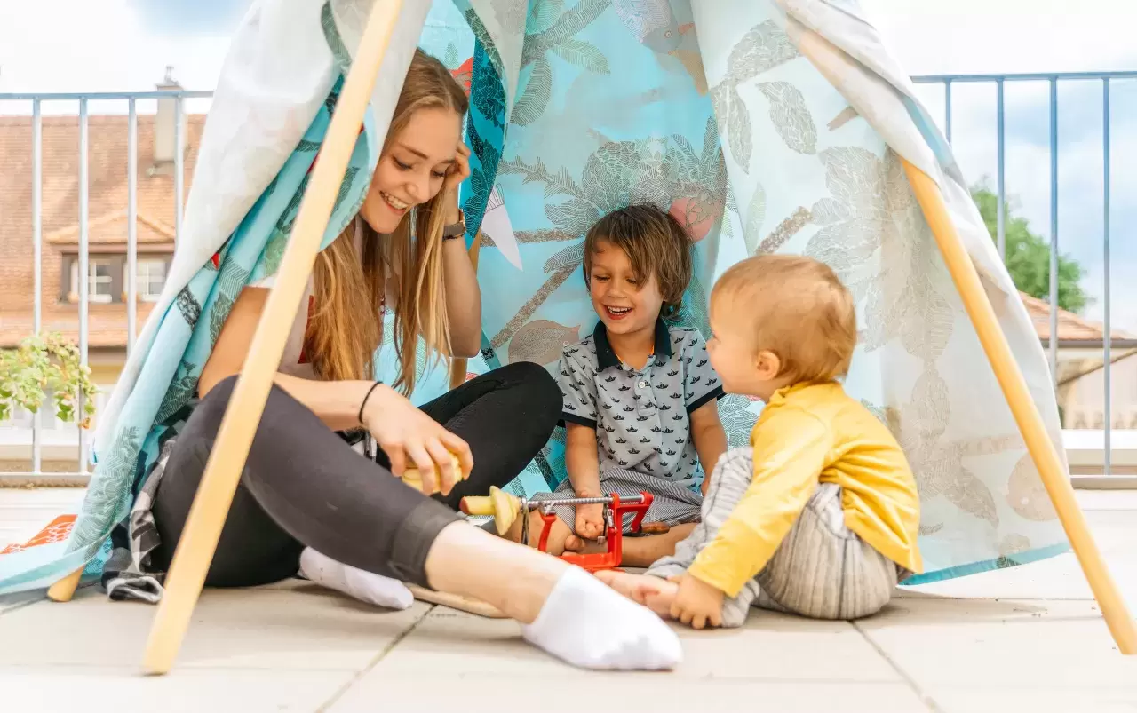 Jeune personne assise et riant avec deux enfants en bas âge dans une tente de jeu.