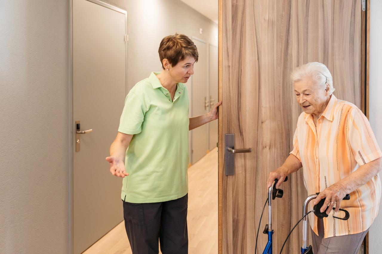 Eine Pflegehelfende hält einer Seniorin am Rollator die Tür auf.