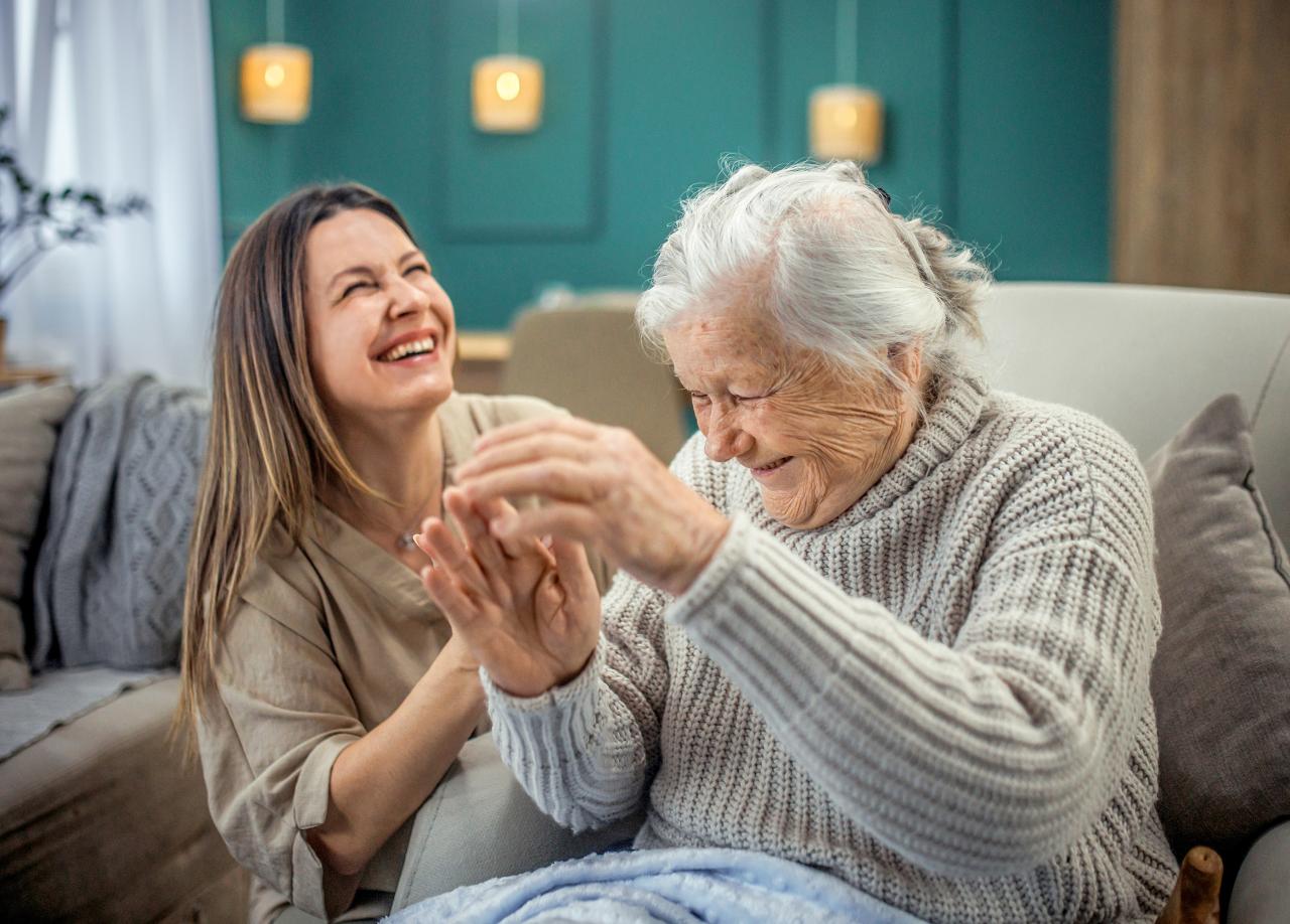 Eine ältere Frau sitzt neben einer jüngeren Frau, die ihre Hand hält. Beide lachen gemeinsam.