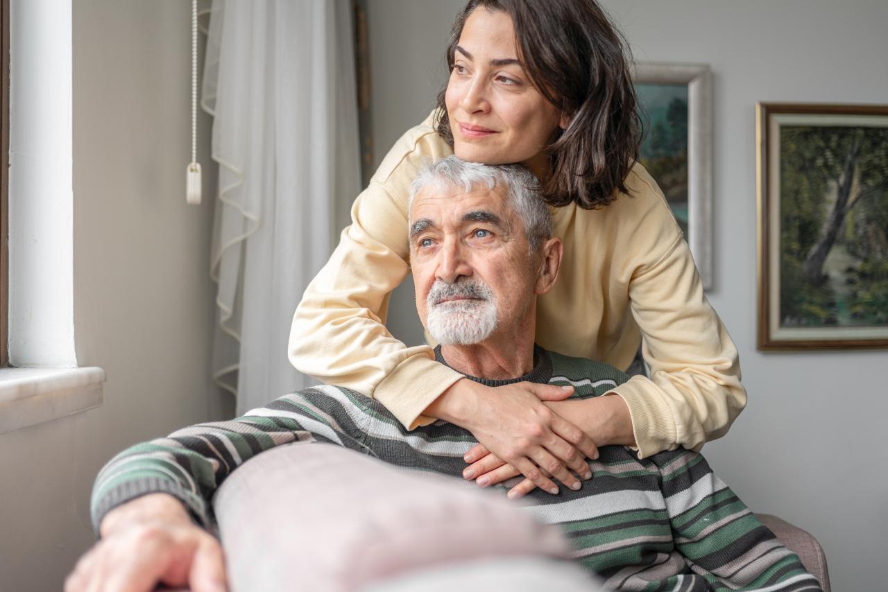 Älterer Mann sitzt in einem Sessel, während eine Frau ihn von hinten umarmt.