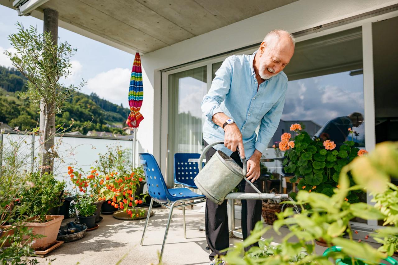 Älterer Mann mit Notrufknopf am Handgelenk giesst zuhause auf dem Balkon die Blumen.