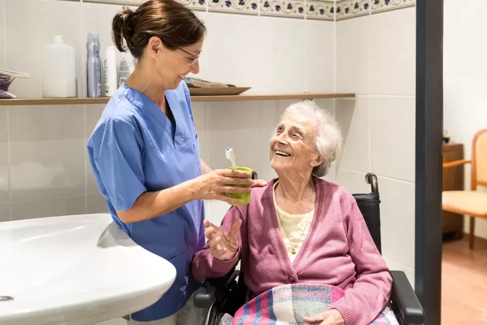 Eine Pflegehelfende reicht einer Seniorin im Rollstuhl den Zahnputzbecher.
