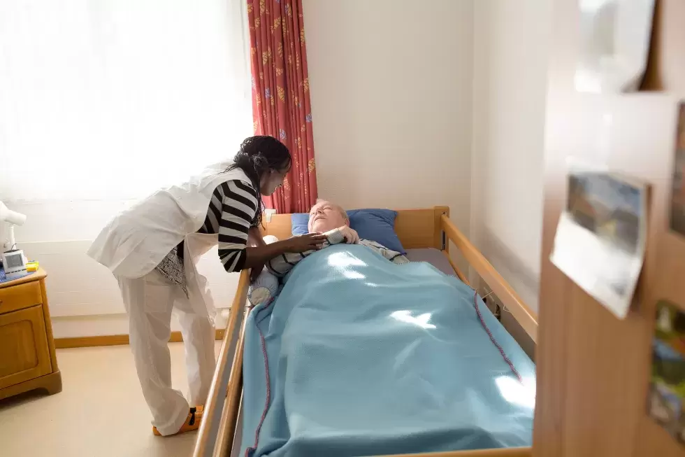Pflegehelfende mit Migrationshintergrund kümmert sich um eine bettlägerige Patientin.