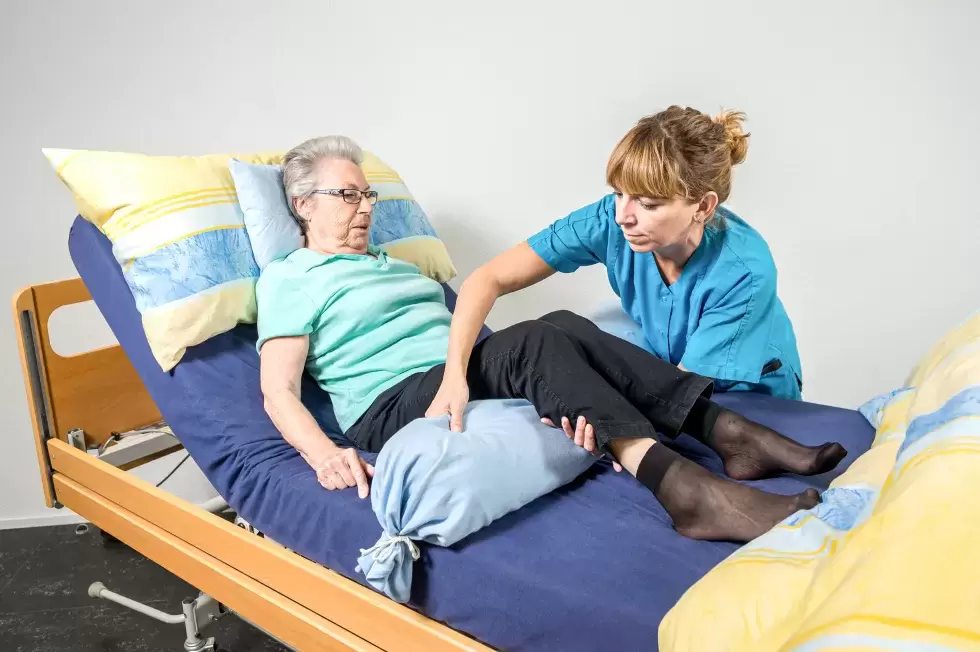 Eine Pflegehelfende unterstützt eine bettlägerige Seniorin dabei, mit Hilfe von einem weiteren Kissen eine komfortablere Position zu finden. 
