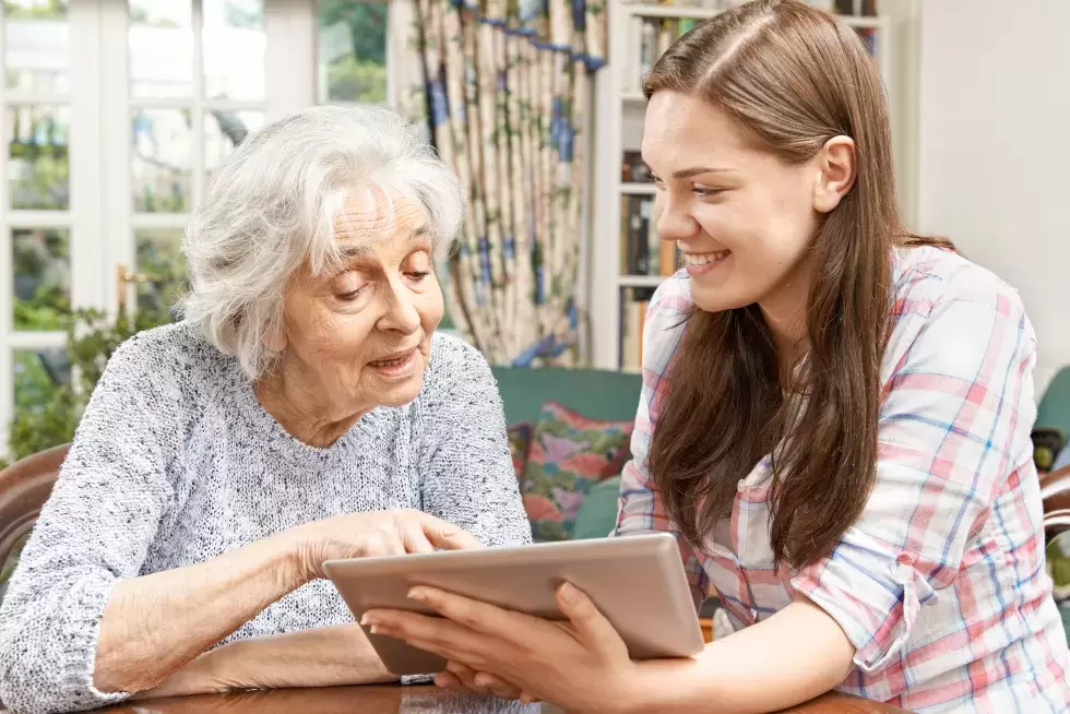 Eine junge Erwachsene hilft einer älteren Dame beim Bedienen eines Tablets.