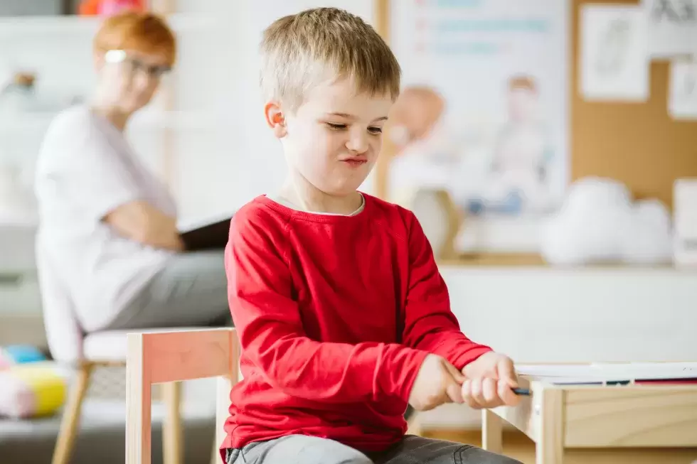 Junge in rotem T-Shirt spielt im Klassenzimmer. Die Sozialarbeiterin im Hintergrund schaut zu.