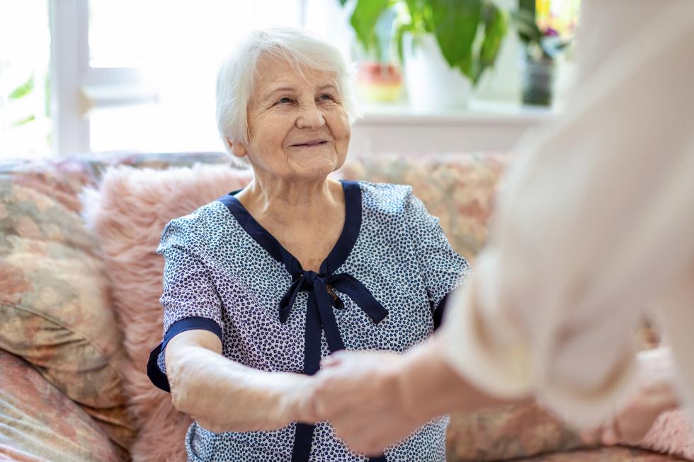 Eine Pflegehelfe reicht einer älteren Frau die Hände.