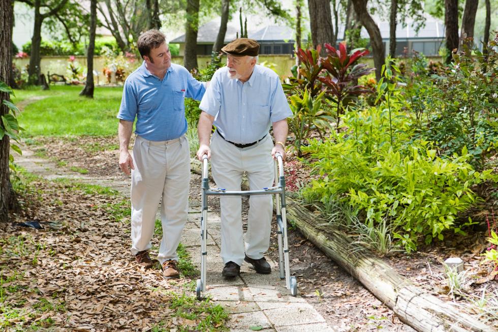 Ein Mann am Rollator wird von einem Pflegehelfenden auf seinem Spaziergang unterstützt.