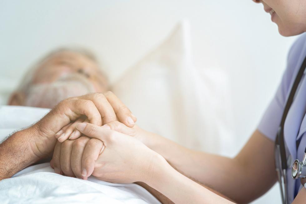 Eine Pflegehelfende hält die Hände eines bettlägerigen Mannes.
