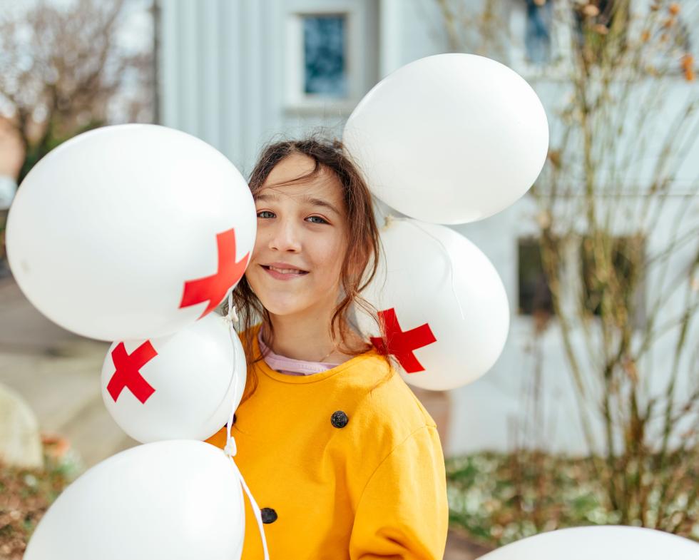 Ein Maedchen lacht in die Kamera, sie ist umgeben von fünf weissen Ballonen mit roten Kreuzen drauf. 