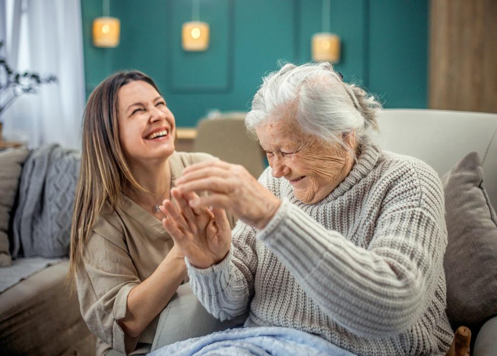 Eine ältere Frau sitzt neben einer jüngeren Frau, die ihre Hand hält. Beide lachen gemeinsam.